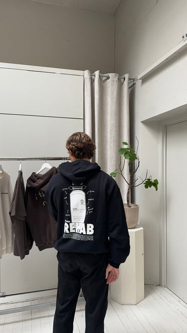 Eine Person mit kurzen braunen Haaren steht in einem Innenraum vor einem Kleiderständer und trägt den DALUMA JOSEPH ATELIER X DALUMA REHAB HOODIE mit einer Rückengrafik und dem Text "REHAB". In der Nähe befindet sich eine Topfpflanze vor einer hellen Wand und Vorhängen.
