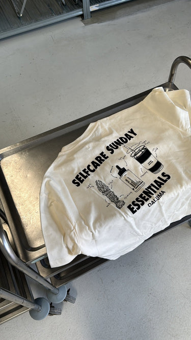 Das JOSEPH ATELIER X DALUMA SELFCARE SUNDAY TEE, ein weißes T-Shirt aus Bio-Baumwolle von DALUMA mit schwarzen Pflanzen- und Selbstpflege-Illustrationen und dem Text SELF-CARE SUNDAY ESSENTIALS DALUMA, wird im nachhaltigen Siebdruckverfahren bedruckt und auf einem Metallwagen in einem hellen Raum ausgestellt.