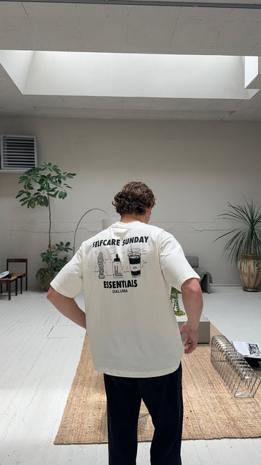 Eine Person mit lockigem Haar, von hinten in einem Innenraum gesehen, trägt das JOSEPH ATELIER X DALUMA SELFCARE SUNDAY TEE von DALUMA mit Illustrationen und Text zur Selbstfürsorge. Im Hintergrund sind ein heller Holzboden, Pflanzen und Möbel zu sehen.