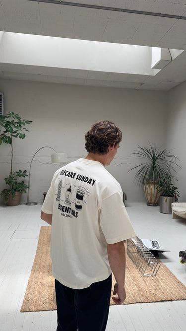 In einem hellen, minimalistischen Raum mit weißen Wänden und moderner Einrichtung trägt eine Person das JOSEPH ATELIER X DALUMA SELFCARE SUNDAY TEE von DALUMA. Der Raum wird durch Topfpflanzen, eine Lampe und einen Teppich akzentuiert.