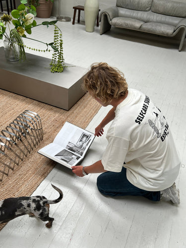 Eine blonde Person, die das DALUMA JOSEPH ATELIER X DALUMA SELFCARE SUNDAY TEE trägt, kniet auf einem weißen Boden und blättert in einem Schwarz-Weiß-Fotobuch, während ein kleiner Hund an modernen Möbeln und Pflanzen vorbeiläuft.