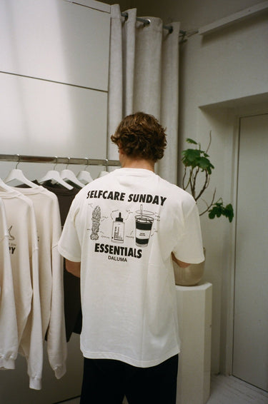 Eine Person mit gewelltem braunem Haar, von hinten gesehen, trägt das DALUMA JOSEPH ATELIER X DALUMA SELFCARE SUNDAY TEE mit nachhaltigen, illustrierten Selbstpflegeartikeln im Siebdruck und "SELFCARE SUNDAY ESSENTIALS DALUMA" auf dem Rücken.