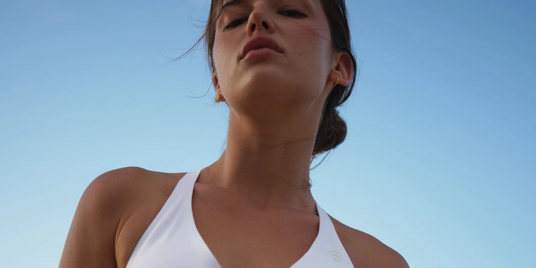 Eine Frau, die einen weißen Badeanzug trägt, blickt mit zurückgebundenen Haaren und einem ruhigen Gesichtsausdruck in die Kamera vor einem klaren blauen Himmel.