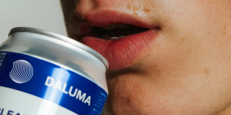 Nahaufnahme der Lippen einer Person, die einen Schluck aus einer silbernen Dose mit einem blau-weißen Etikett mit der Aufschrift "DALUMA" neben einem weißen, runden Logo nimmt.
