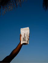 Eine Hand hält einen weißen Beutel mit der Aufschrift PERFORMANCE DUO KAKAO von DALUMA vor einem klaren blauen Himmel hoch, über dem Palmenblätter hängen - ideal für den Muskelaufbau mit pflanzlichem Eiweiß.