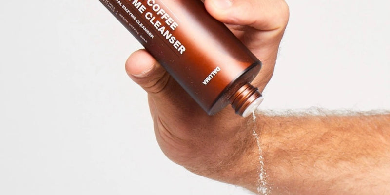 Eine Hand gießt ein flüssiges Reinigungsmittel aus einer braunen Flasche mit der Aufschrift Coffee Crème Cleanser auf den Rücken einer anderen Hand, vor einem schlichten weißen Hintergrund.