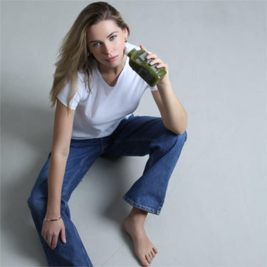 Eine Frau mit langen blonden Haaren, weißem T-Shirt und blauen Jeans, sitzt barfuß mit einem Glas grünen Saft aus ihrer DALUMA GRÜNE SAFTKUR PLUS - 3 TAGE und schaut in die Kamera.
