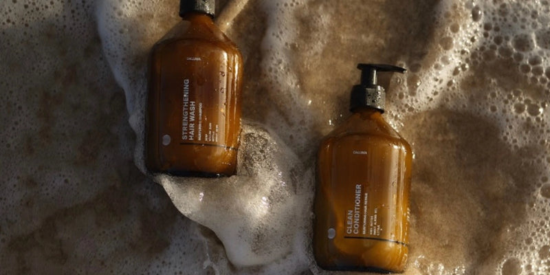 Zwei braune Pumpflaschen mit den Aufschriften "Hair Wash" und "Conditioner" liegen auf dem nassen Sand, teilweise bedeckt von den schäumenden Wellen des Meeres.