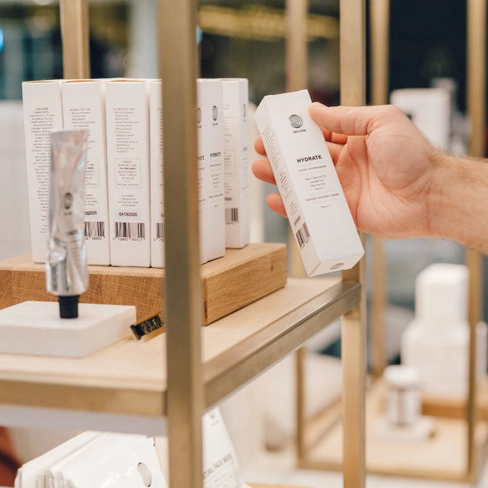 Die Hand einer Person nimmt eine Schachtel mit der Aufschrift HYDRATE aus einem Regal mit verschiedenen Hautpflegeprodukten und Schachteln, die in einem Geschäft ordentlich angeordnet sind.