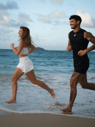 Eine Frau und ein Mann joggen barfuß am Ufer entlang, lächelnd in der Meeresbrise unter einem teilweise bewölkten Himmel. Als Mitglieder des DALUMA HEALTH CLUB PRO MEMBERSHIP kommen sie in den Genuss exklusiver Veranstaltungen zur Förderung von Wohlbefinden und sportlicher Vitalität.