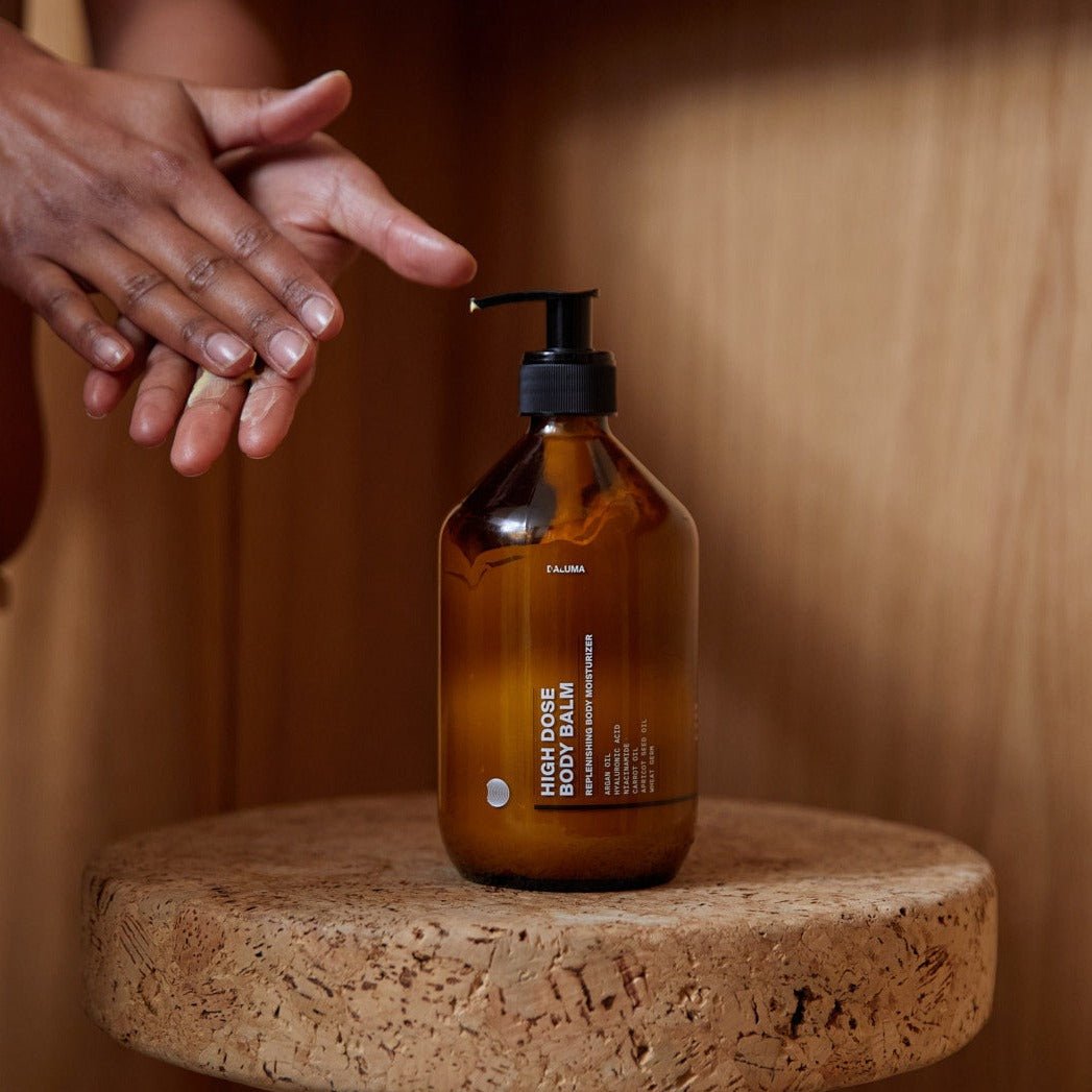 Eine Person reibt ihre Hände in der Nähe einer bernsteinfarbenen DALUMA HIGH DOSE BODY BALM-Pumpflasche auf einer Korkoberfläche, die vor einem hölzernen Hintergrund für intensive Feuchtigkeitspflege steht.