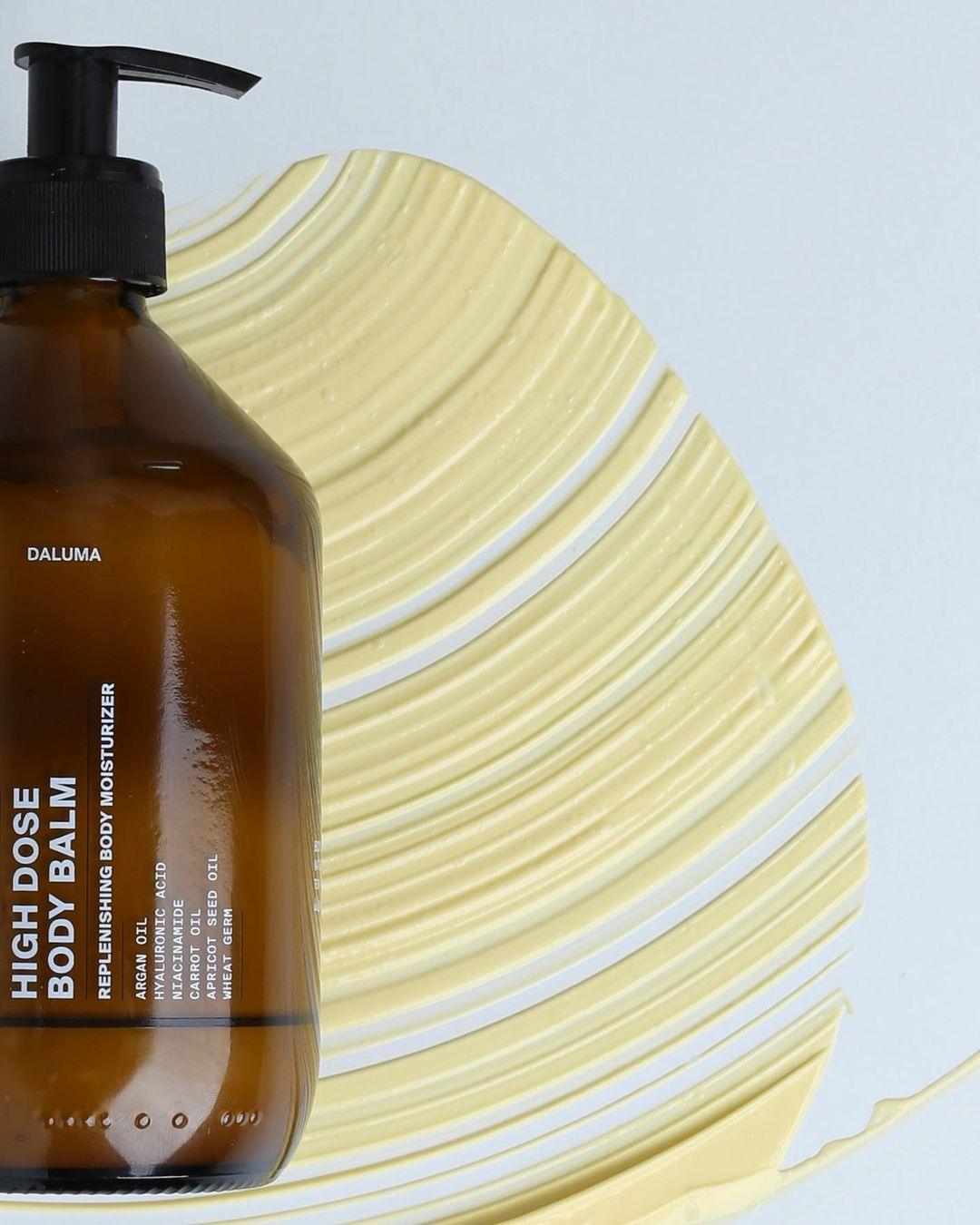 Eine braune Pumpflasche mit DALUMA HIGH DOSE BODY BALM steht auf einer hellen Fläche, neben der ein großzügiger Klecks gelber Balsam - reich an Arganöl für intensive Feuchtigkeitspflege und Anti-Aging-Effekte - kunstvoll verteilt ist.