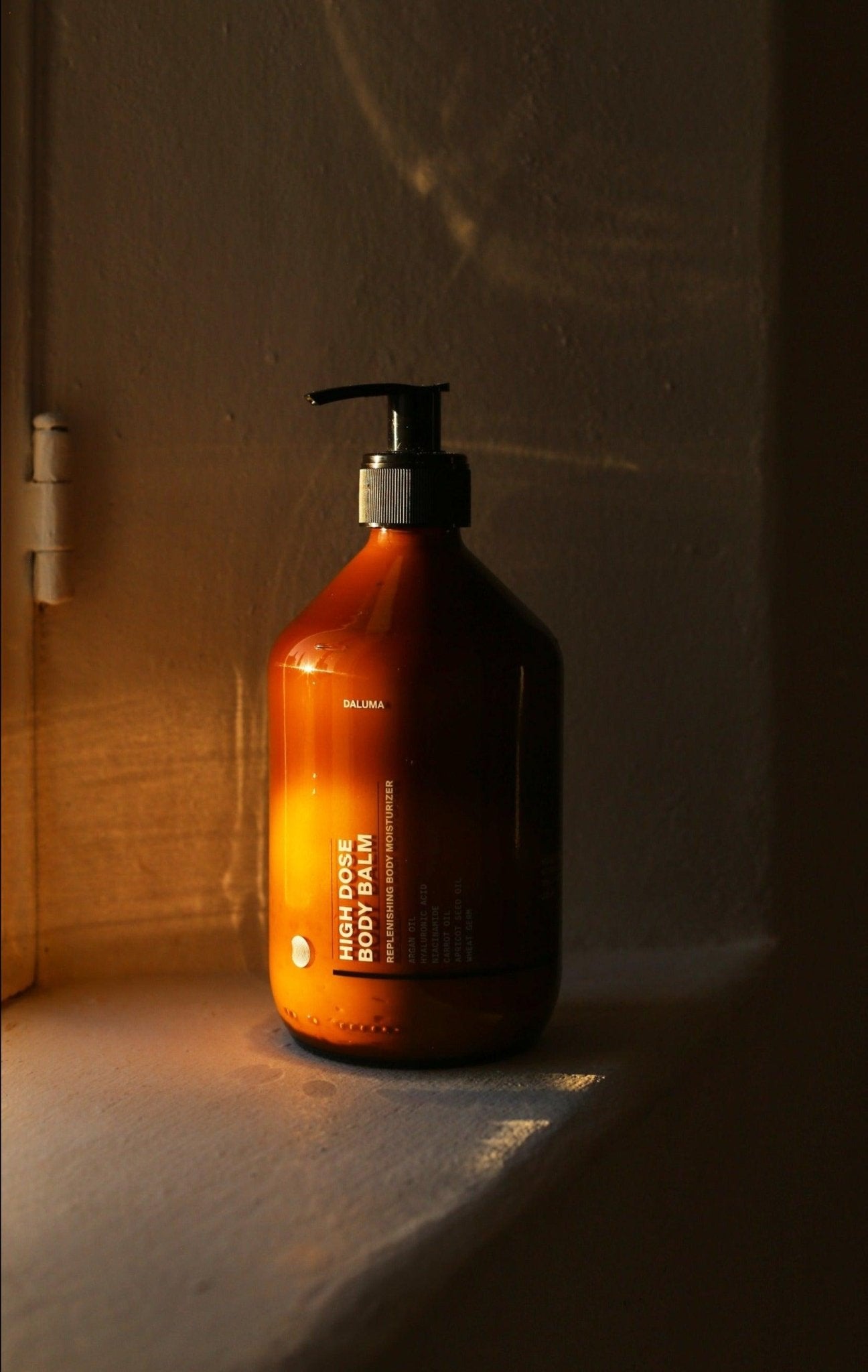 Eine goldene, bernsteinfarbene Pumpflasche mit DALUMA HIGH DOSE BODY BALM steht auf einer weißen Fensterbank, die in warmes Sonnenlicht getaucht ist, das durch das nahe gelegene Fenster fällt.