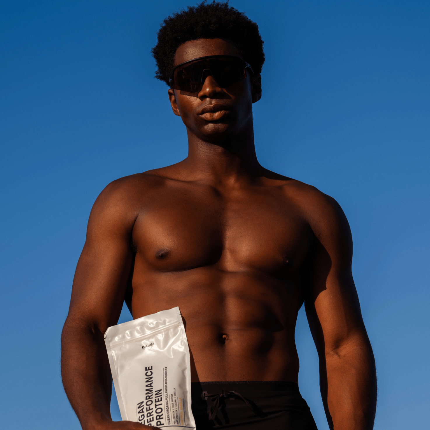 Ein durchtrainierter Mann ohne Hemd und mit schwarzer Sonnenbrille steht vor einem strahlend blauen Himmel und hält den ENERGY & STRENGTH DUO-Beutel von DALUMA in der Hand, der mit pflanzlichem Eiweiß und veganen BCAAs für optimalen Muskelaufbau sorgt.