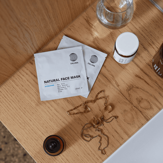 Zwei DALUMA HYDRATING ESSENTIALS Gesichtsmasken, ein kleines Glas, eine goldene Halskette, ein Glasbecher und eine braune Flasche stehen auf einer hellen Holzfläche neben einem weißen Waschbecken.