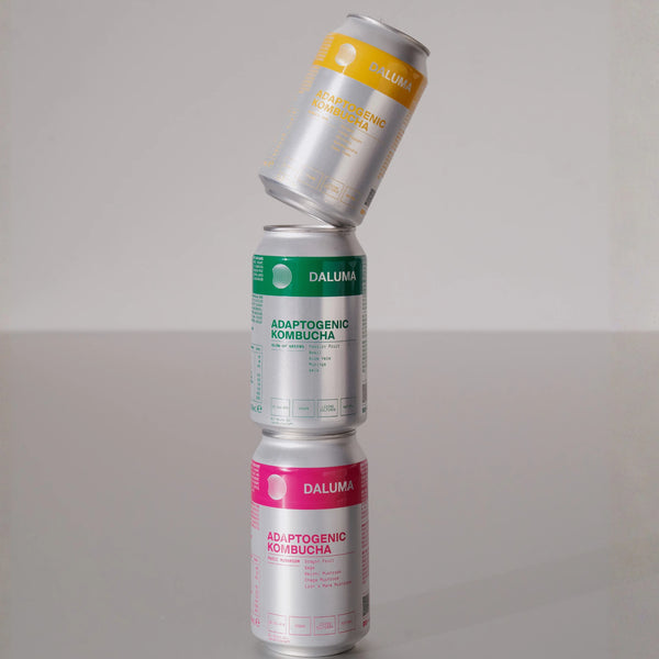 Drei Dosen Daluma Adaptogenic Kombucha vertikal gestapelt, wobei eine gelbe Dose auf einer grünen und einer rosa Dose vor einem schlichten hellgrauen Hintergrund balanciert.
