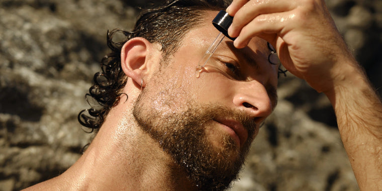 Ein Mann mit nassem Haar und Bart trägt mit einer Pipette Gesichtsserum auf seine Wange auf, während er draußen vor einem felsigen Hintergrund steht.