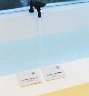 Eine Badewanne füllt sich mit Wasser, und zwei Beutel DALUMA 2x MASK BUNDLE liegen auf dem Rand. Im Hintergrund ist ein Fenster mit Grünpflanzen zu sehen.