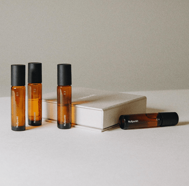 Die Nullpunkt COMPLETE COLLECTION von Nullpunkt zeigt vier Braunglas-Rollerflaschen mit schwarzen Verschlüssen, die neben einem geschlossenen beigen Buch auf weißem Untergrund und neutralem Hintergrund stehen; drei Flaschen stehen aufrecht, eine liegt auf der Seite.