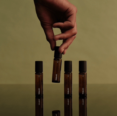 Eine Hand hält eine bernsteinfarbene Rollflasche der NULLPUNKT COMPLETE COLLECTION von Nullpunkt über einer reflektierenden Oberfläche. Drei passende Flaschen und minimalistische Etiketten stehen vor einem Hintergrund in gedämpftem Grün für einen eleganten, natürlichen Look.