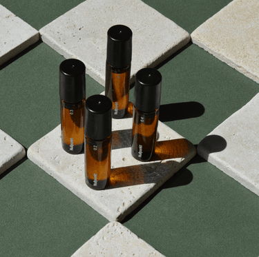 Vier kleine Braunglasflaschen aus der NULLPUNKT COMPLETE COLLECTION von Nullpunkt, jeweils mit schwarzem Verschluss, sind auf cremefarbenen Kacheln vor grünem Hintergrund angeordnet, die Schatten werfen und ein auffälliges geometrisches Muster bilden.