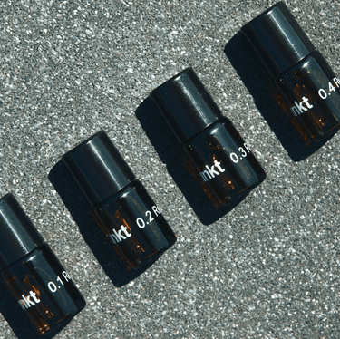 Vier kleine Braunglasflaschen mit schwarzen Deckeln, die mit "0.1", "0.2", "0.3" und "0.4" beschriftet sind, aus dem Nullpunkt NULLPUNKT DISCOVERY SET sind diagonal auf einer gesprenkelten grauen Fläche angeordnet und werfen Schatten nach rechts.