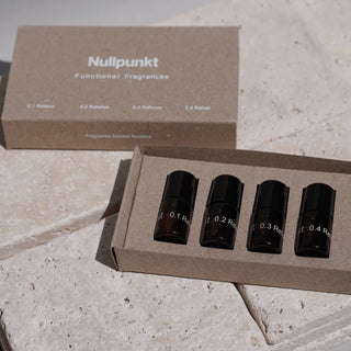 Eine Pappschachtel mit der Aufschrift Nullpunkt Functional Fragrances ist geöffnet und enthält vier kleine dunkle Glasflaschen, die jeweils mit einer Nummer und einem Wort beschriftet sind. Der Karton steht auf einer beigen Steinfläche.