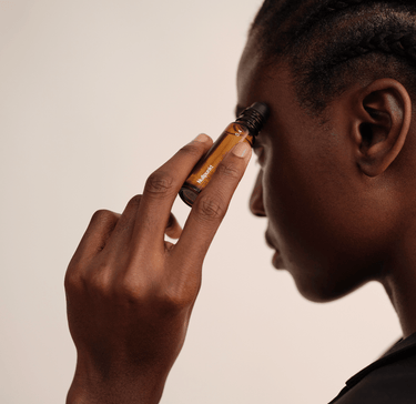 Eine Person mit geflochtenen Haaren trägt die Rollerflasche Nullpunkt NULLPUNKT GRADE 0.4 REFUEL sanft an der Schläfe auf und hält sie vor einem neutralen Hintergrund dicht an ihr Gesicht.
