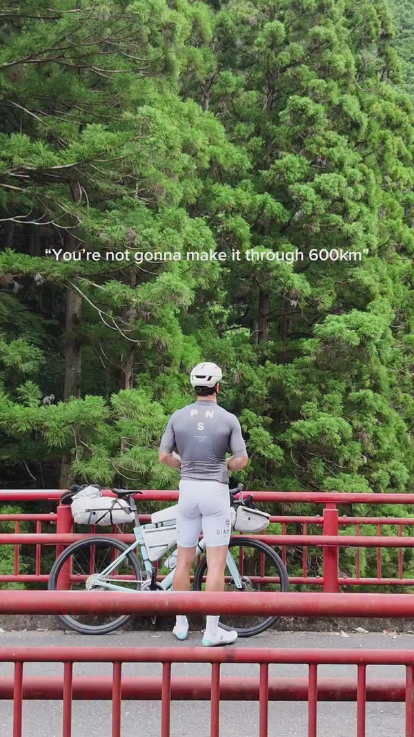 Ein Radfahrer in grauer Kleidung steht mit einem beladenen Fahrrad auf einer roten Brücke, vor dichten grünen Bäumen. Der Text darüber lautet: "Du wirst es nicht schaffen, 600 km zu fahren".