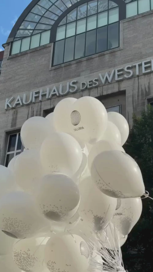 Vor dem Kaufhaus des Westens schwebt an einem sonnigen Tag ein Bündel weißer Luftballons mit Logos. Der Name des Gebäudes ist in großen Buchstaben über dem Eingang zu lesen.