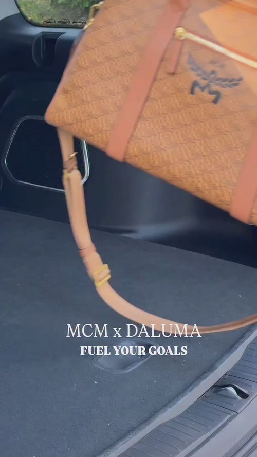 Ein hellbrauner MCM x Daluma Seesack wird in den Kofferraum eines Autos gelegt. Der Text auf dem Bild lautet: MCM x DALUMA FUEL YOUR GOALS.