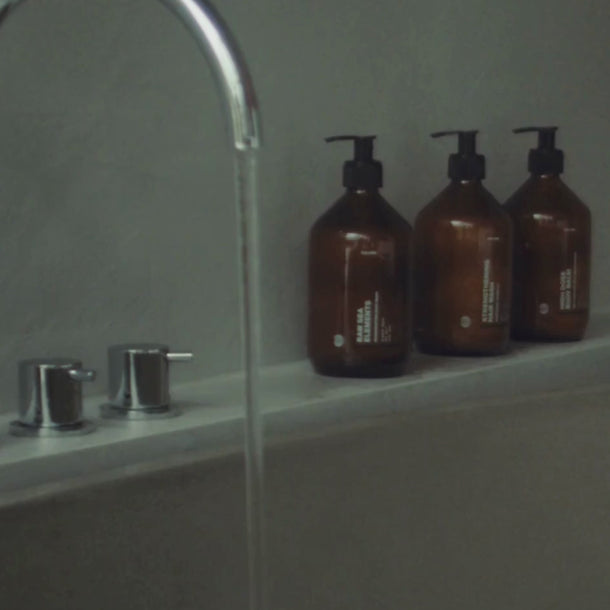 Eine Nahaufnahme eines Badewannenhahns mit fließendem Wasser neben drei braunen Daluma HAIR CARE DUO Pumpflaschen auf dem Wannenrand vor einer grauen Wand.