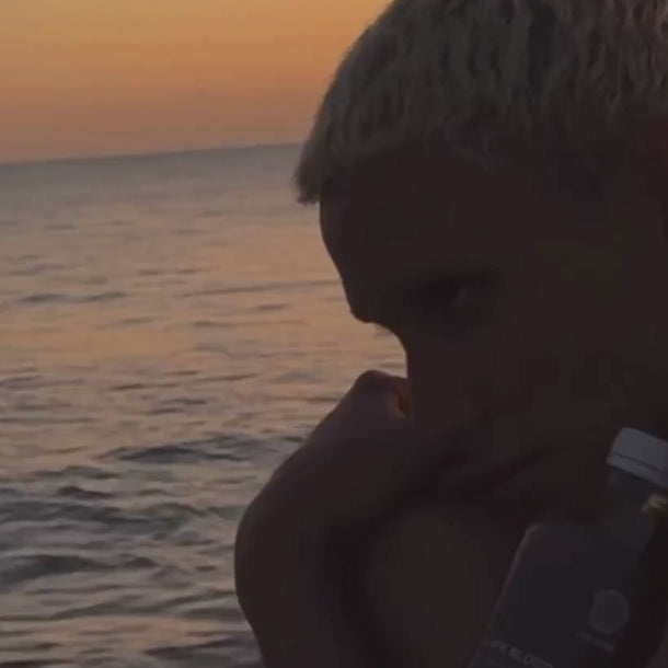 Eine Person mit kurzem, hellem Haar sitzt bei Sonnenuntergang am Meer und hält ein Daluma ALKALISCHE SAFTKUR - 3 TAGE Getränk in der Hand, während sich im ruhigen Wasser der orangefarbene Himmel spiegelt.