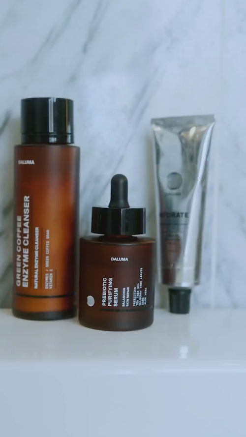 Drei Hautpflegeprodukte auf einer weißen Badezimmeroberfläche: eine große braune Flasche mit der Aufschrift "Enzyme Cleanser", ein braunes Tropffläschchen mit der Aufschrift "Probiotic Serum" und eine silberne Tube vor einem Marmorwandhintergrund.