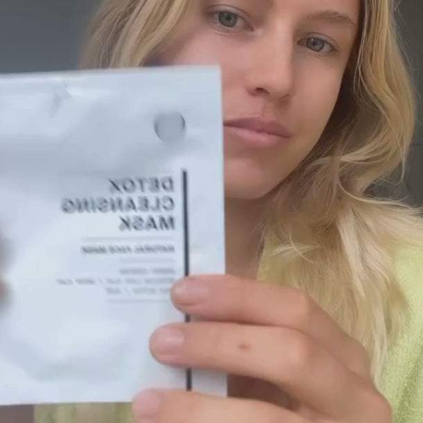 Eine Frau mit langen blonden Haaren hält eine Packung mit der Aufschrift Detox Cleansing Mask in die Kamera, wobei ihr Gesicht teilweise verdeckt wird. Sie scheint in einem Haus zu sein und trägt ein helles Oberteil.