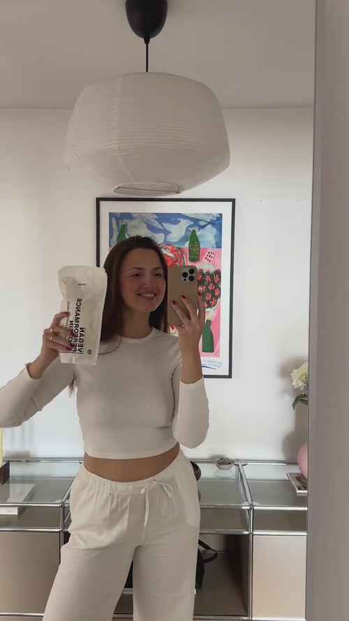 Eine Frau in einem weißen Crop-Top und einer Jogginghose macht ein Selfie im Spiegel, während sie lächelt und ein weißes Paket hält. Im Hintergrund sind eine moderne Hängelampe und ein gerahmtes Kunstwerk zu sehen.