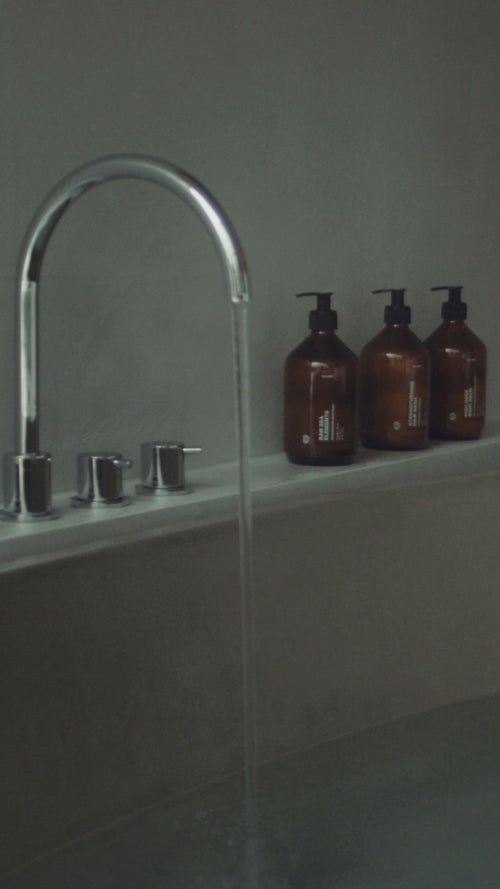 Eine Nahaufnahme einer Badewanne, die sich mit Wasser aus einem verchromten Wasserhahn füllt. Die braunen Pumpflaschen von Daluma CLEANSE COUPLE stehen auf dem Wannenrand vor einer grauen Wand und schaffen eine minimalistische und heitere Szene.