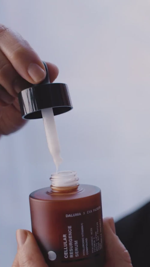 Eine Hand hält einen Tropfer mit weißem Serum über einer offenen braunen Flasche mit der Aufschrift "CELLULAR RESURGENCE SERUM" von Daluma x Eva Padberg vor einem schlichten hellen Hintergrund.