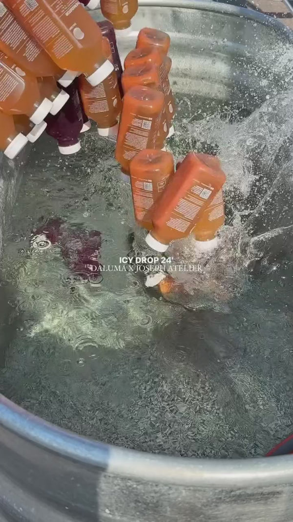 Mehrere orangefarbene und lilafarbene Flaschen werden in eine große, mit Wasser gefüllte Metallwanne geworfen, wodurch ein Spritzer entsteht. Der Text über dem Bild lautet: ICY DROP 24 DALUMAX JOSEPH ATELIER.