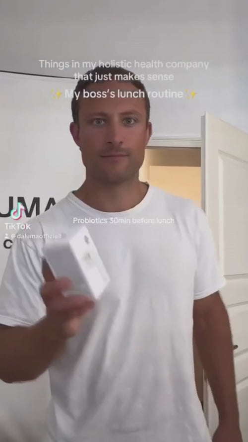 Ein Mann in einem weißen T-Shirt steht in einem Raum, hält eine weiße Schachtel und schaut in die Kamera. Der Text auf dem Bild erwähnt ein ganzheitliches Gesundheitsunternehmen, die Mittagsroutine des Chefs und Probiotika 30 Minuten vor dem Mittagessen.