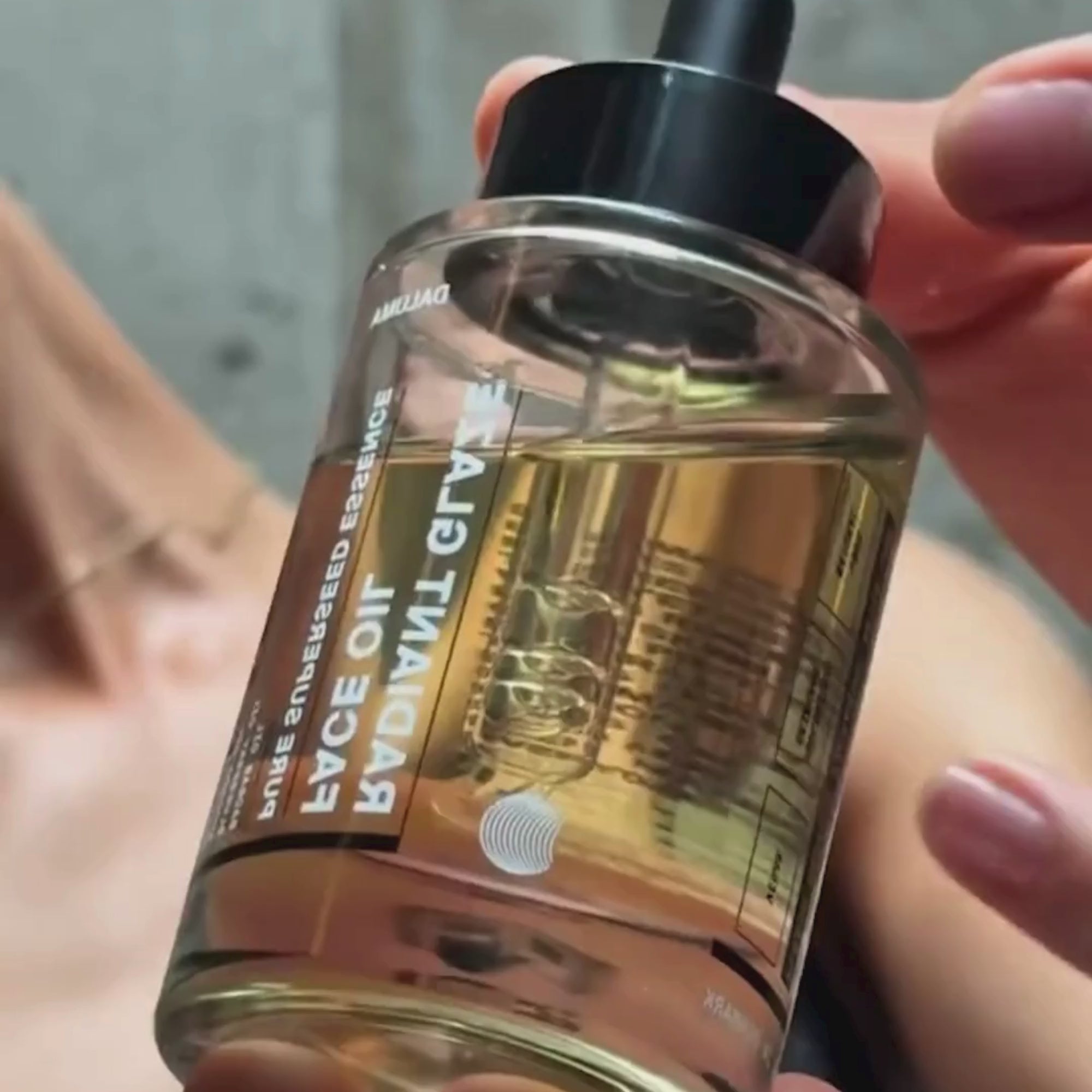 Eine Hand hält eine Klarglasflasche mit einer bernsteinfarbenen Flüssigkeit und einem Tropfer mit der Aufschrift GRAVITY FACE OIL in fetten Buchstaben in der Nähe des Gesichts einer Person im verschwommenen Hintergrund.