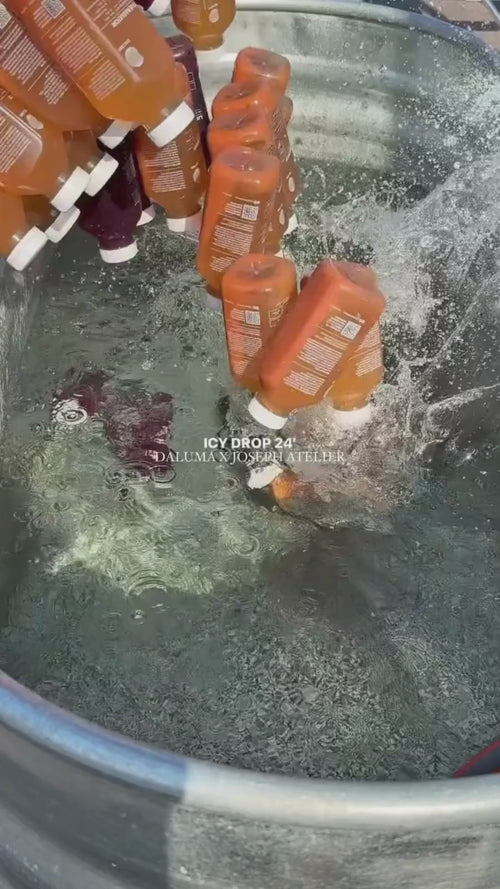 Mehrere orangefarbene Plastikflaschen mit der Aufschrift HEALTH CLUB PRO MEMBERSHIP von Daluma werden in eine große Metallwanne mit eiskaltem Wasser geworfen und plätschern darin. In der Mitte erscheint der Text ICY DROP 24 DALUMA X JOSEPH HADLEDER.
