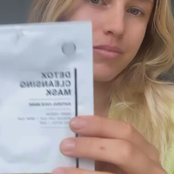 Eine Person mit langem blondem Haar hält eine Packung mit der Aufschrift Detox Cleansing Mask in die Kamera, wodurch ihr Gesicht teilweise verdeckt wird. Der Hintergrund ist neutral und unscharf.