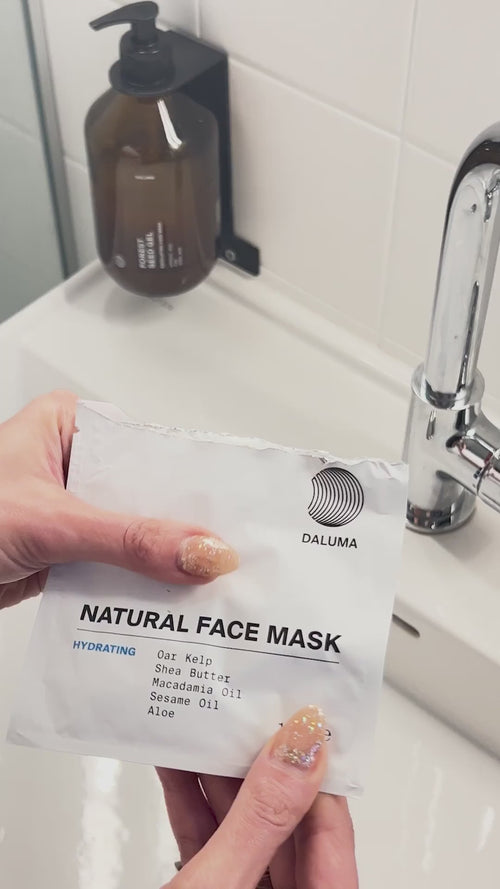 Eine Hand mit weiß lackierten Nägeln hält eine teilweise geöffnete weiße Packung Daluma DEEP HYDRATION FEUCHTIGKEITSMASKE, in der eine cremefarbene beigefarbene Maske zum Vorschein kommt.