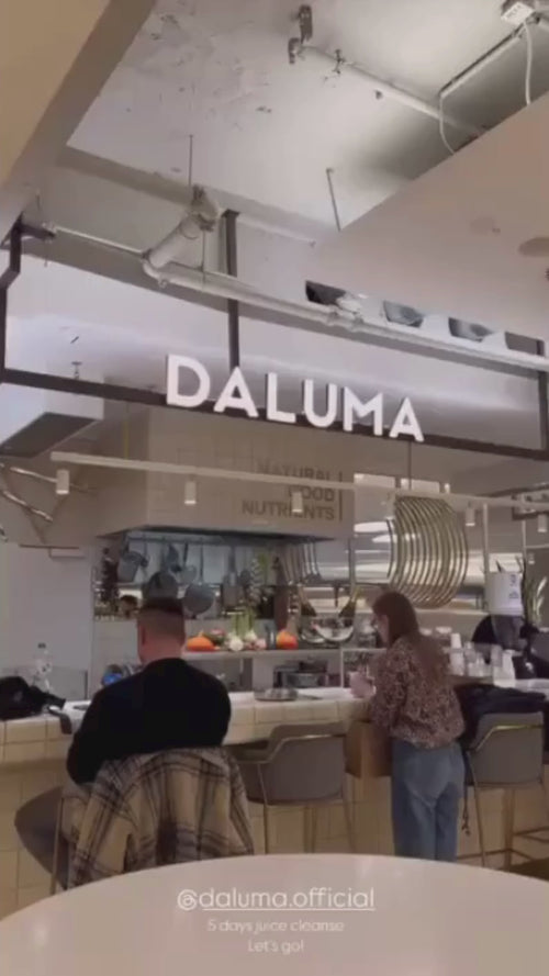 Ein modernes Café mit einem Schild mit der Aufschrift DALUMA über dem Tresen. Zwei Personen sitzen auf Hockern an der Theke, die eine trägt eine karierte Jacke, die andere hat lange Haare. Man sieht runde Regale und Küchengeräte.