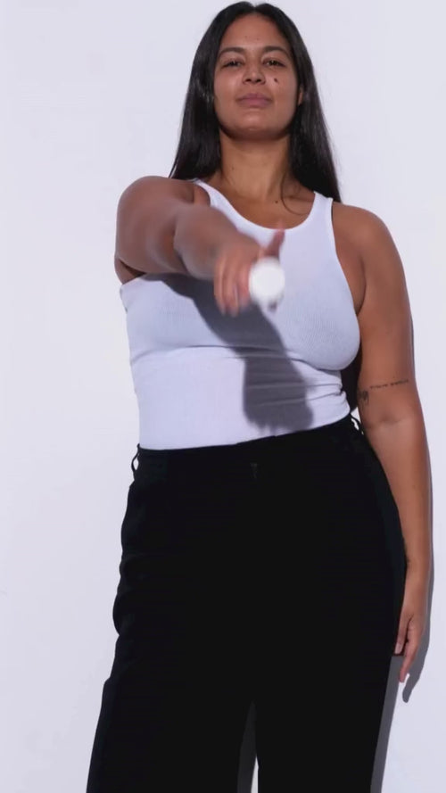 Eine Frau mit langen dunklen Haaren in einem weißen ärmellosen Oberteil und einer schwarzen Hose steht vor einem weißen Hintergrund und hält selbstbewusst die BETTER GUT SAFTKUR 3 TAGE Saftkur von Daluma in die Kamera.