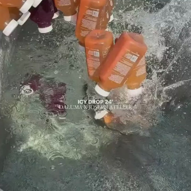 Mehrere orangefarbene Flaschen mit weißen Verschlüssen, die mit Daluma DAILY HEALTH PACKAGE beschriftet sind, werden in klares Wasser geworfen, wodurch Spritzer und Blasen entstehen. Der weiße Text auf dem Bild lautet: "ICY DROP 24" und "DALUMA x JOSEPH ATELIER".