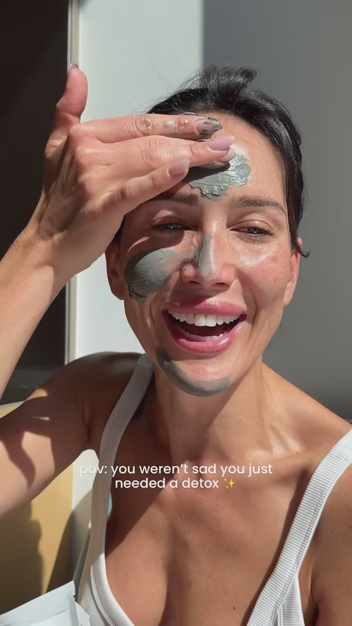 Eine lächelnde Frau, die sich eine graue Gesichtsmaske auf Stirn und Nase legt und ein weißes Tanktop trägt. Das Sonnenlicht beleuchtet ihr Gesicht. Der Text auf dem Bild lautet: pov: you werent sad you just needed a detox ✨.