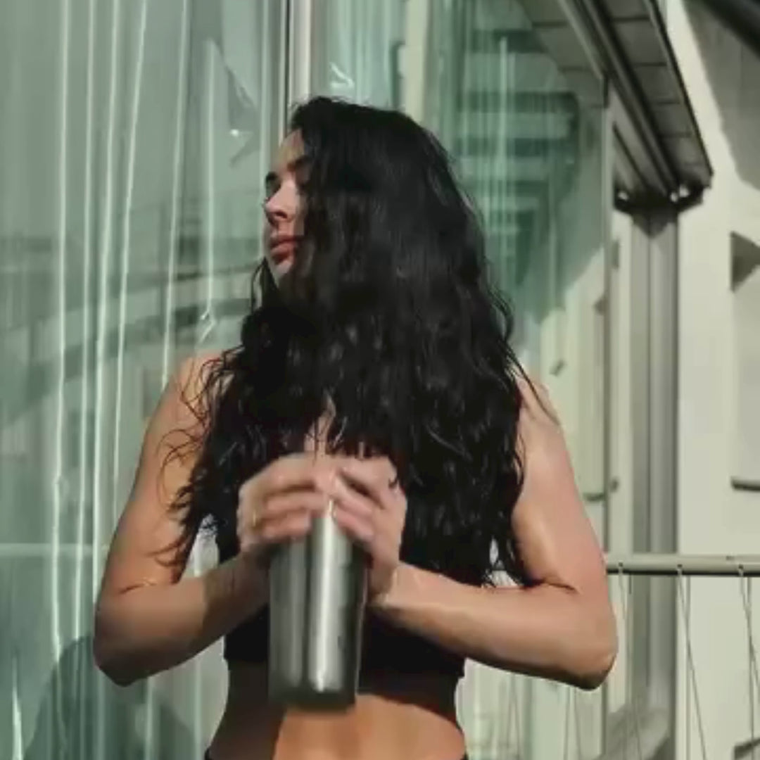 Eine Frau mit langen dunklen Haaren steht im Sonnenlicht auf einem Balkon, trägt einen schwarzen Sport-BH und hält mit beiden Händen eine metallene Shakerflasche. Sie blickt zur Seite, und hinter ihr sind Glasscheiben zu sehen.