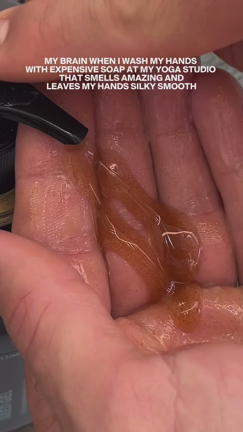 Eine Nahaufnahme zeigt Dalumas CLEANSE COUPLE, eine bernsteinfarbene Flüssigseife, die auf eine Handfläche aufgetragen wird. Der eingeblendete Text scherzt darüber, dass man sich diese luxuriöse, duftende Seife nach dem Yoga gönnen sollte und dass sie die Hände seidenweich macht.